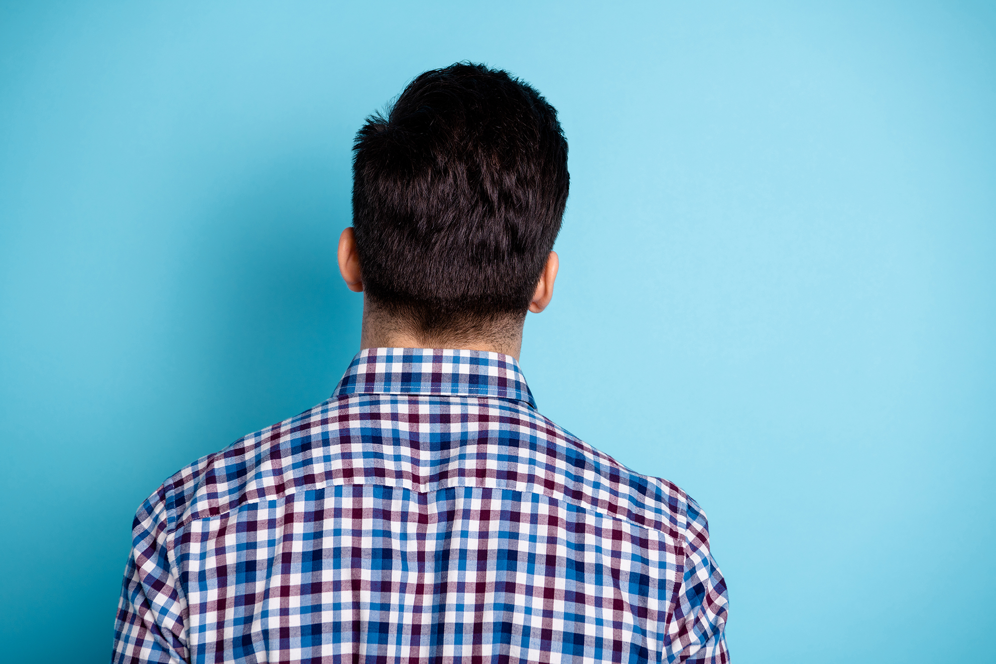 Henkilö seisoo turkoosia seinää vasten selin kameraan. Hänellä on ruutupaita ja lyhyet tummat hiukset.